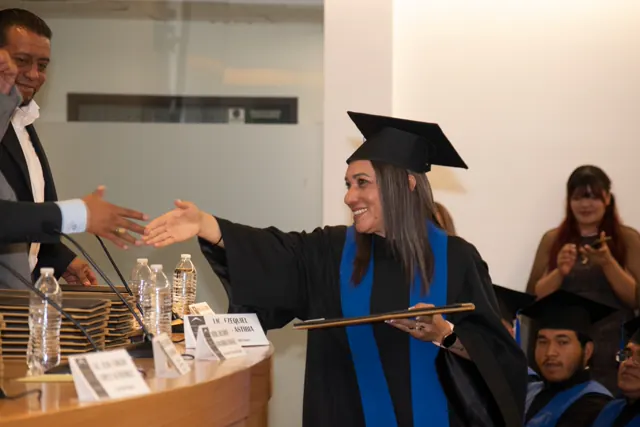 Graduada sonriente con toga negra y estola azul saluda mientras sostiene su carpeta de titulación.