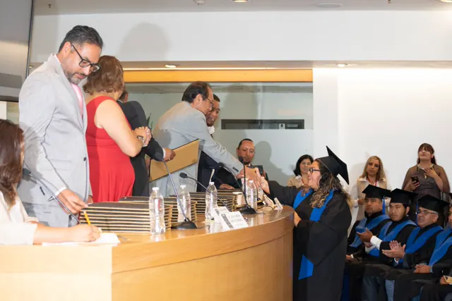 Graduada con toga y birrete recibe su carpeta ante la mesa de entrega de títulos.