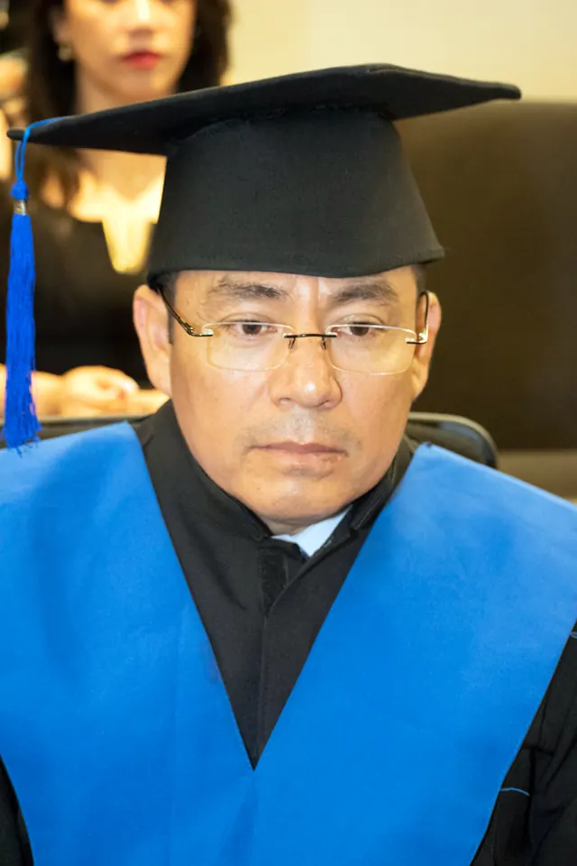 Graduado con lentes, birrete negro y estola azul escucha durante la ceremonia.