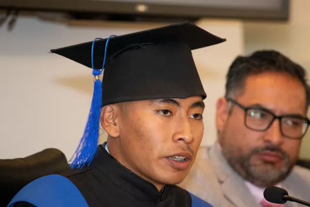 Joven graduado con birrete y estola azul habla ante un micrófono.