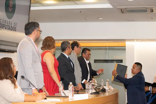 Egresado con traje azul recibe su carpeta de titulación frente a los presentadores.