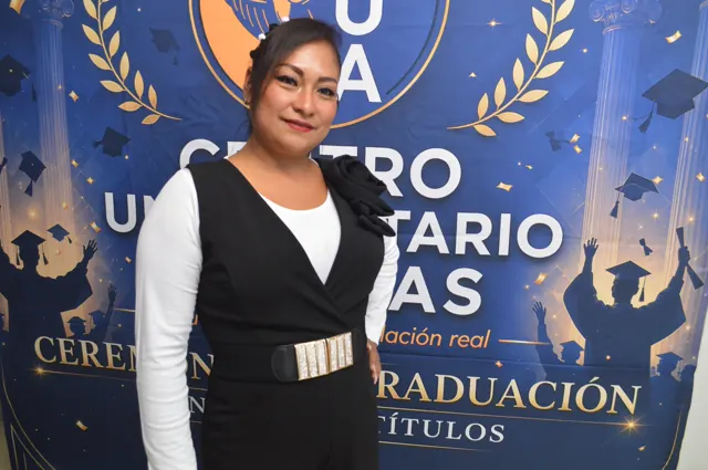 Participante con conjunto negro y blusa blanca posa ante el fondo institucional de la graduación.