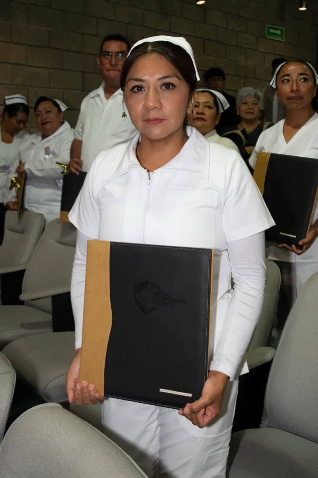Egresada de enfermería con cofia y uniforme blanco muestra su carpeta. Graduación del 14 de junio de 2025.