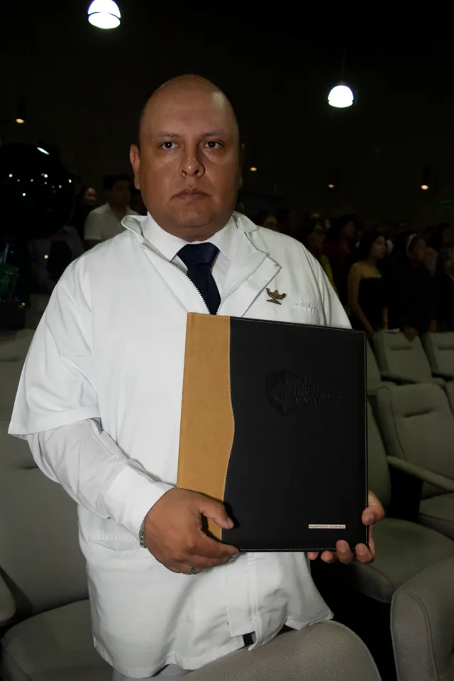 Egresado con bata blanca y corbata sostiene su carpeta de graduación. Graduación del 14 de junio de 2025.